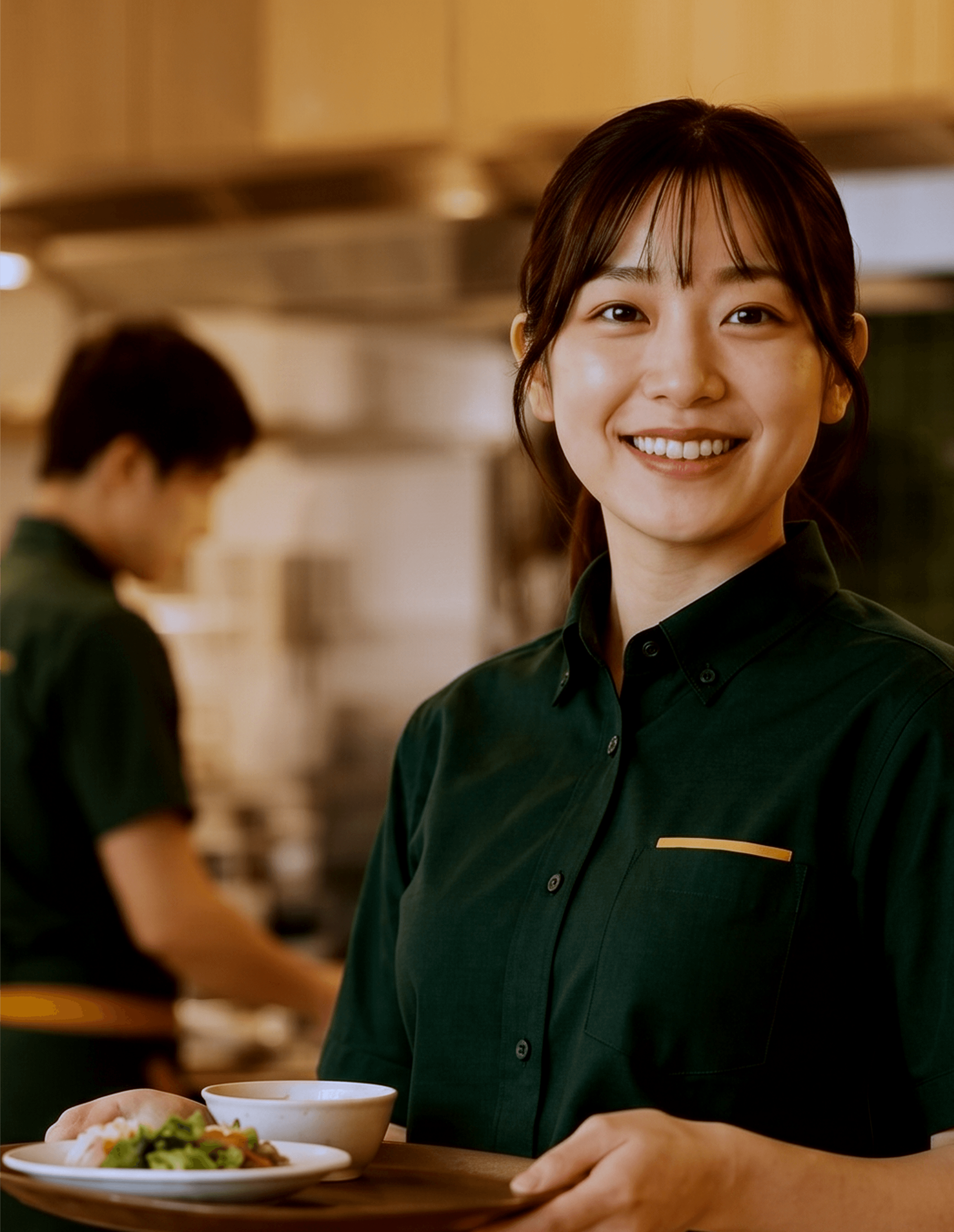 制服姿の飲食店スタッフが料理を運びながら笑顔を見せる厨房の様子